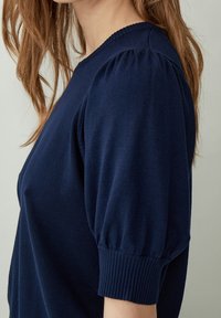 Mörkblå stickad tröja med puffärmar, ribbade muddar och rund halsringning, som visar en slät textur.