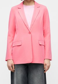 Femme portant un blazer rose vif à un seul bouton sur un haut rose clair et un pantalon large gris foncé, coupé à mi-cuisse.