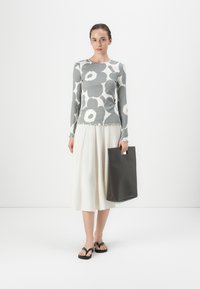 Grå bluse med langt erme og blomstermønster, med volanger i bunnen, kombinert med et hvitt skjørt og svarte flip-flops, mens hun holder en mørkegrå tote-veske.