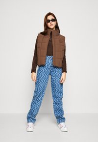 Gilet imbottito marrone a quadratini sopra un maglione marrone, abbinato a jeans a gamba larga blu a fantasia e sneakers bianche. Occhiali da sole neri completano il look.