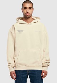 Kræmfarvet hættetrøje med frontlomme, ribbede manchetter og minimalistiske tekstdetaljer. Stoffet ser blødt og struktureret ud, afslappet pasform.