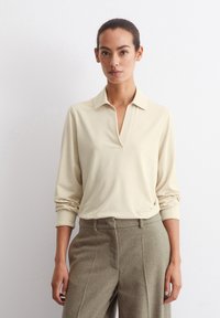 Beige Langarm-Polohemd mit Kragen, das einen V-Ausschnitt und einen lockeren Schnitt aufweist, kombiniert mit grauen, hochtaillierten Tweed-Hosen.