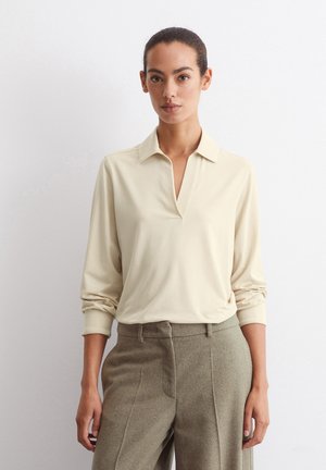 Beige Langarm-Polohemd mit Kragen, das einen V-Ausschnitt und einen lockeren Schnitt aufweist, kombiniert mit grauen, hochtaillierten Tweed-Hosen.