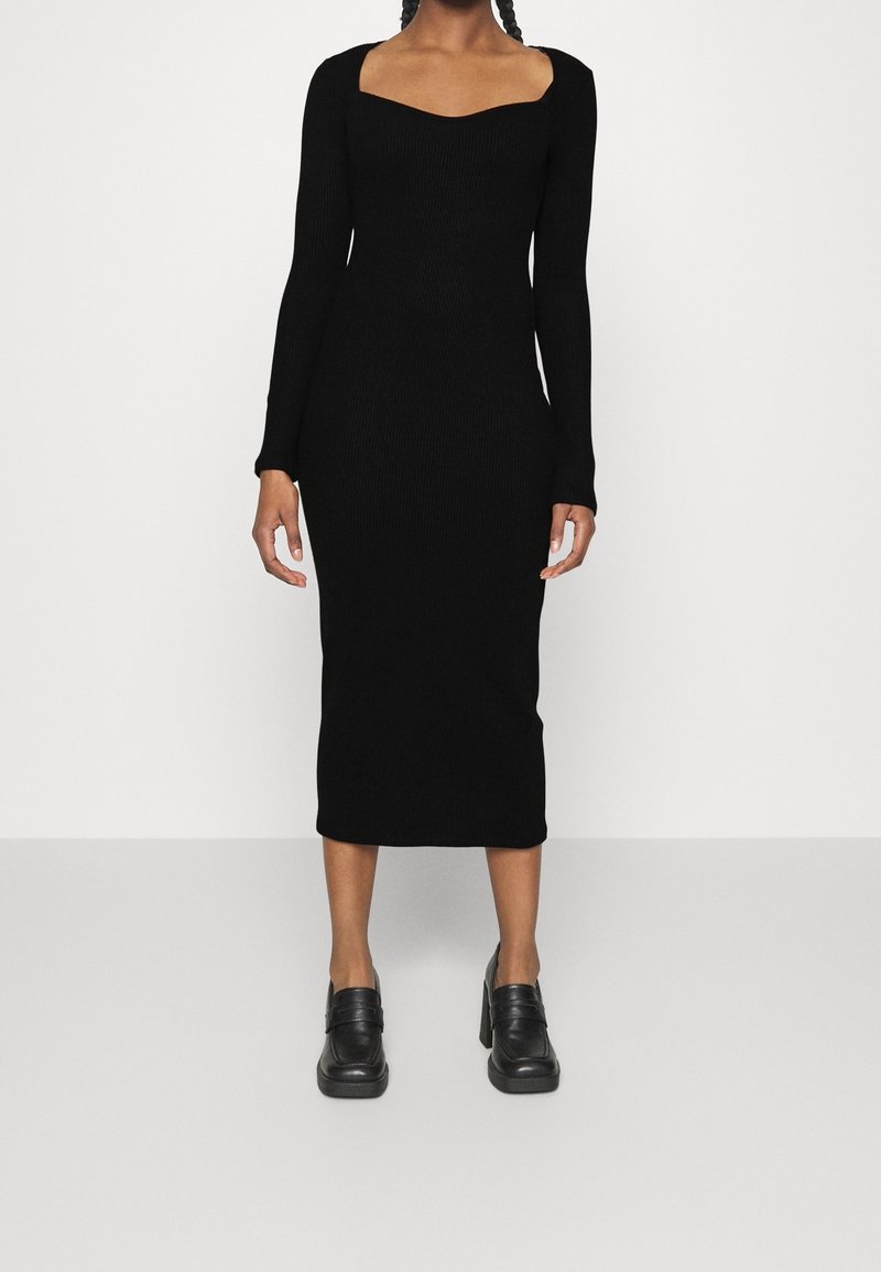 Robe noire côtelée à manches longues avec un décolleté carré et une longueur mi-mollet, associée à des mocassins noirs à talons. Texture lisse, coupe ajustée.