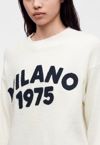 Jeune personne portant un pull blanc avec un texte noir texturé indiquant "MILANO 1975" à l'avant sur un fond uni.