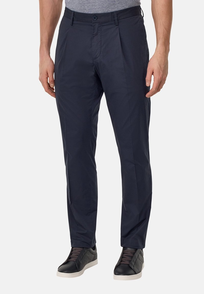 Homme portant un pantalon bleu marine foncé slim fit avec des plis, associé à des baskets noires avec des semelles blanches et des lacets gris, debout devant un fond uni.