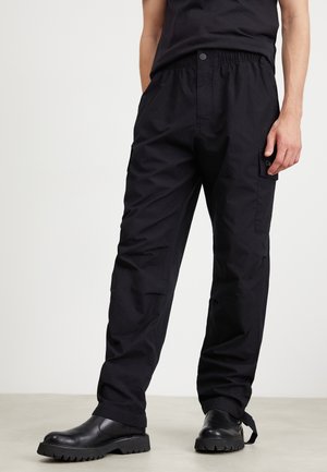 Mann trägt schwarze Cargo-Hose mit Seitentasche und schwarze Lederstiefel, steht auf hellem Boden vor weißem Hintergrund.