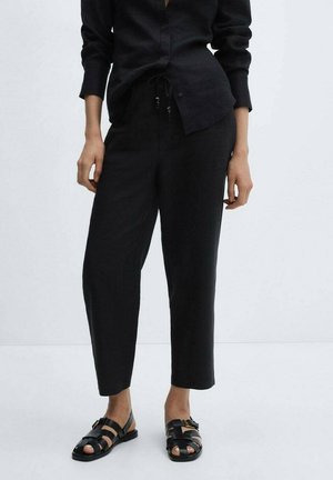 Personne portant un pantalon noir fuselé avec taille à cordon, une chemise noire boutonnée et des sandales noires à boucle, debout sur un fond clair.