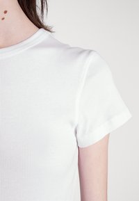 Detailní záběr na rameno a krk osoby oblečené v bílém krátkém tričku s žebrováním, na krku jsou tři malé hnědé pihy.