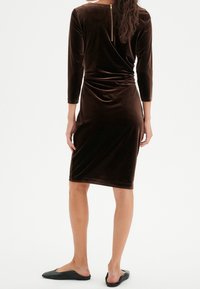 Brun sammetsklänning med 3/4 ärmar, sidofåll och dragkedja bak. Kombinerad med svarta platta skor på en modell mot en neutral bakgrund.