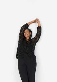 Top in pizzo nero con maniche lunghe caratterizzato da motivi floreali, abbinato a pantaloni neri. Il top ha una vestibilità ampia e dettagli elaborati.