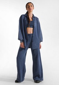 Outfit in denim composto da una camicia blu oversize, un crop top e pantaloni a gamba larga; texture morbida con bottoni e dettagli con cordini.