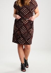 Brun mönstrad klänning med korta ärmar, med geometriska vita mönster. Bärs med ett svart bälte och svarta öppna ankelstövlar.