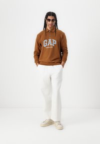 Brun hettegenser med "GAP ATHLETIC" logo, hvite vide bukser og beige sneakers. Modellen har solbriller på og har flettet hår.