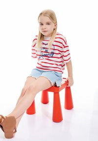 Rood-wit gestreepte T-shirt met blauwe tekst, gecombineerd met licht denim shorts met rafelige randen. Zit op een felrode kruk.