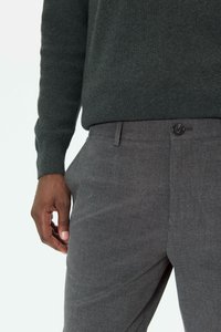 Mand iført mørkegrøn sweater og grå bukser, med venstre hånd afslappet ved siden mod en lys baggrund.