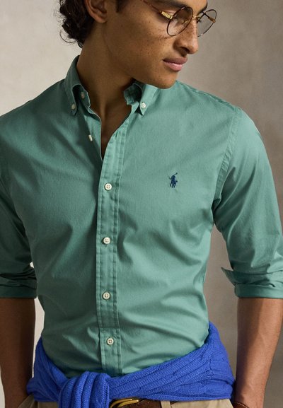 Jeune homme portant une chemise verte boutonnée et un pull bleu noué autour de la taille, regardant vers le bas et sur le côté, avec des lunettes rondes.