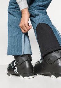 Calças de esqui em veludo cotelê azul com pormenores em tecido preto. Apresenta zíperes nas pernas, um ajuste descontraído e são usadas com botas de esqui pretas.