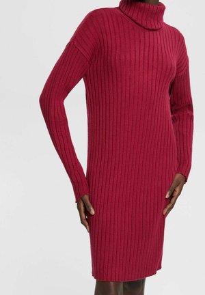 Červené rebrované pletené šaty s dlouhými rukávy a vysokým rolákem. Látka má hladkou strukturu a přiléhavý střih, končící nad kolenem.