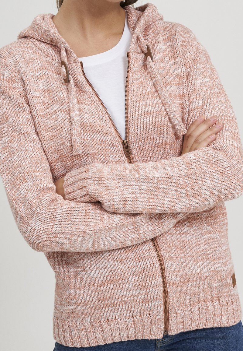 Person med en rosa och vit stickad zip-up hoodie med dragsko och en vit skjorta under, armarna i kors.