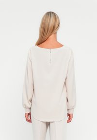 Μακριμάνικο μπλούζα σε ανοιχτό μπεζ ύφασμα με στρογγυλή λαιμόκοψη και φερμουάρ στο πίσω μέρος. Έχει μαζεμένα μανίκια και χαλαρή εφαρμογή.