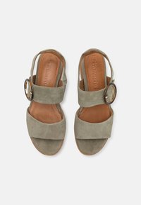 Suede sandaler i oliven grønn med to brede stropper. Har en rund metallspenne som dekorasjon og en lys lærsåle. Flatt, åpen bakdesign.