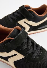 Chaussures de sport noires en suède et mesh, avec un accent marron clair, des lacets et une sangle velcro, conçues dans un style vintage.