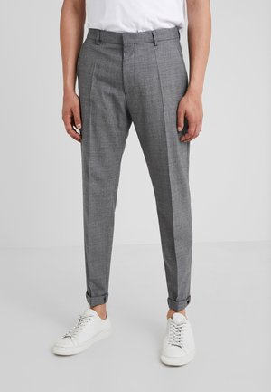 Person trägt schmal geschnittene graue Anzughose mit Umschlägen, kombiniert mit weißen Sneakers und einem weißen Hemd, steht vor einem einfarbigen Hintergrund.