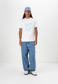 Tricou alb cu un design grafic abstract albastru, asortat cu blugi baggy ușor albaștri și adidași albi. O șapcă albastră completează ținuta.