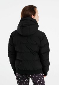 Schwarze Daunenjacke mit Kapuze, ausgestattet mit horizontalen, gesteppten Abschnitten und elastischen Bündchen. Kombiniert mit gemusterten Blumenhosen.