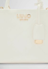 Sac à main en simili cuir blanc avec quincaillerie dorée, logo embossé, forme rectangulaire et étiquette détachable. Détails de surface texturée.