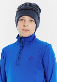 Felpa blu con zip e colletto alto, con logo sul petto, abbinata a un berretto navy e nero con una fascia decorata.