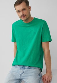 Zielona bawełniana koszulka z krótkim rękawem, okrągłym dekoltem i subtelnym logo. Noszona z jasnoniebieskimi dżinsami.