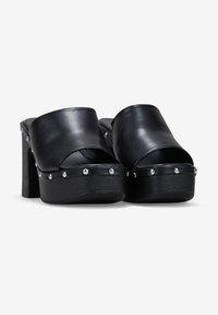 Mules de plataforma de cuero negro con la punta abierta, un tacón ancho y remaches de metal a lo largo del borde de la suela. Textura suave con un acabado brillante.