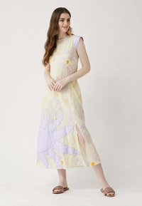 Maxi mekko vaalean keltaisena, jossa on violetteja ja oransseja kukkakuoseja. Siinä on sivusivu ja lyhyet lila hihat. Yhdistetty ruskeisiin litteisiin sandaaleihin.
