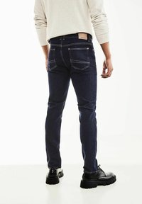 Pantalones vaqueros de mezclilla azul oscuro con corte ajustado, con costuras blancas, dos bolsillos traseros y una etiqueta marrón en la cintura. Llevados con botas negras.