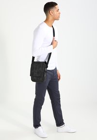 Borsa a messenger nera con una texture in pelle liscia, tracolla regolabile e chiusura a patta. Modello che indossa una camicia bianca a maniche lunghe e pantaloni scuri.