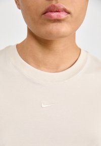 Beige bomullströja med rund halsringning och en liten, broderad vit Nike-logga på framsidan. Slät yta och figurnära design.