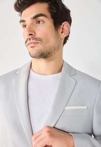 Hellgrauer Blazer aus strukturiertem Stoff, mit einem weißen Taschentuch, getragen über einem schlichten weißen T-Shirt.