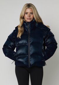 Giacca puffer navy con colletto alto, chiusura a zip e dettagli con bottoni a scatto. Finitura lucida, sezioni trapuntate e dettagli con orlo regolabile.