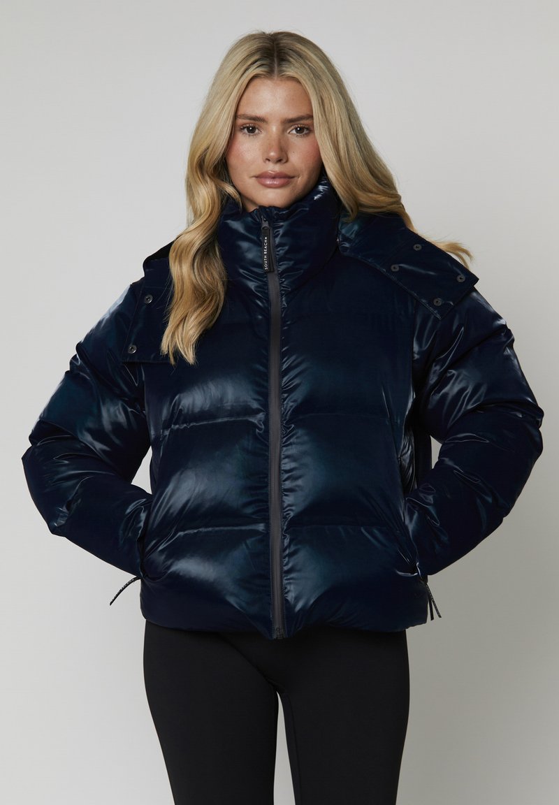 Giacca puffer navy con colletto alto, chiusura a zip e dettagli con bottoni a scatto. Finitura lucida, sezioni trapuntate e dettagli con orlo regolabile.