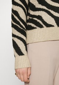 Juostuotasis megztinis su tekstūruotu trikotažu juodos ir beige spalvos. Ribbinti rankogaliai ir apatinė dalis, dėvimi su šviesiomis kelnėmis.