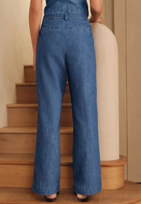 Mėlynos spalvos vidutinio atspalvio platėjantys džinsiniai kelnės su tekstūruotu paviršiumi, elastinga juosmens juosta ir dviem kišenėmis gale, išdėstyti ant medinių laiptų.