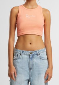 Žena oblečená v broskvovém zkráceném tílku s bílým logem a světlých modrých džínách s vysokým pasem na jednobarevném pozadí.