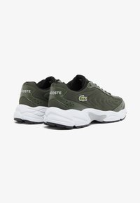 Scarpe sportive verde oliva con tessuto texturizzato, suola intermedia bianca e suola esterna nera. Presentano un logo ricamato sul tallone e dettagli ventilati.