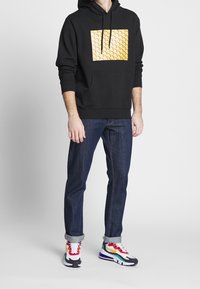 Black hoodie with a textured yellow square design, paired with dark blue jeans and multicolored athletic sneakers.