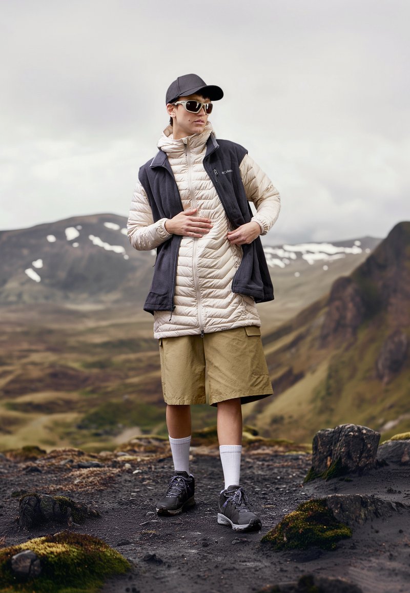 Persoon in een beige jack, grijze vest, tan shorts, witte sokken, zwarte schoenen, pet en zonnebril, staande op rotsachtig terrein met een bergachtig decor.
