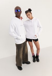 To modeller står med hvite hettegensere med frontlommer mot en nøytral bakgrunn. Den mannlige modellen har grått hår og solbriller; den kvinnelige modellen har på seg shorts.
