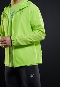 Veste légère vert lime avec capuche, poignets élastiques et poches zippées, associée à des leggings noirs avec un logo blanc.