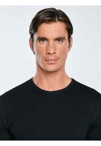 Mann mit ordentlich gekämmtem dunklem Haar und blauen Augen, der ein schwarzes T-Shirt mit Rundhalsausschnitt trägt und vor einem schlichten, hellen Hintergrund nach vorn blickt.